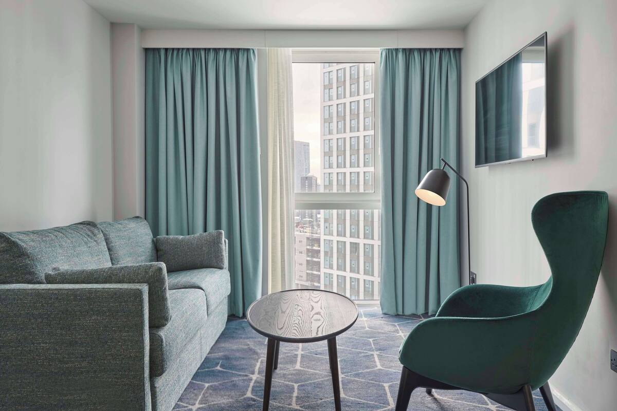 Ein modernes Hotelzimmer mit einem grauen Sofa, einem runden Tisch, einem grünen Sessel und einem großen Fenster mit Vorhängen.