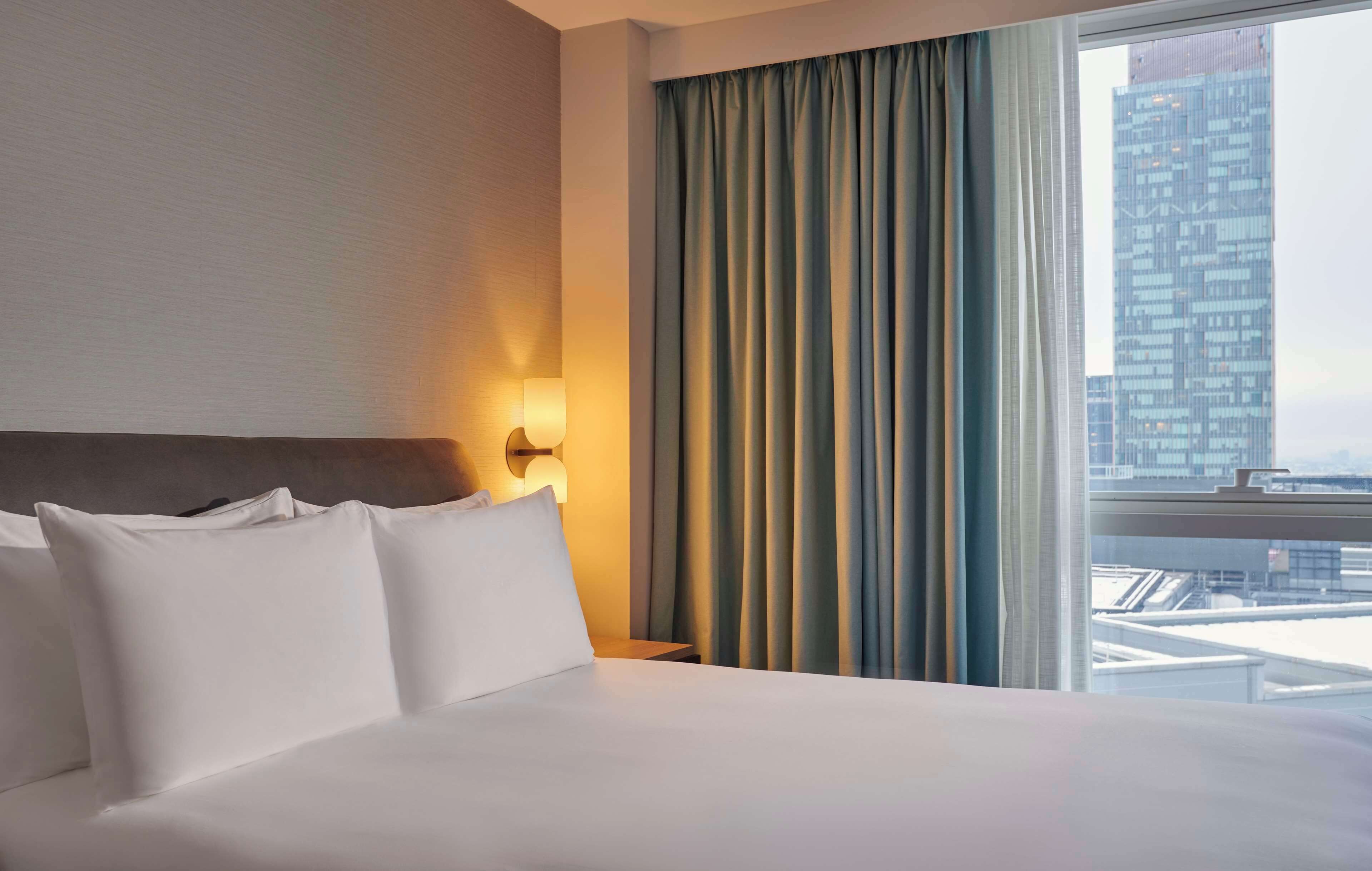 Un cuarto de hotel con una cama grande, una mesita de noche con lámpara y vistas a la ciudad a través de una ventana con cortinas.