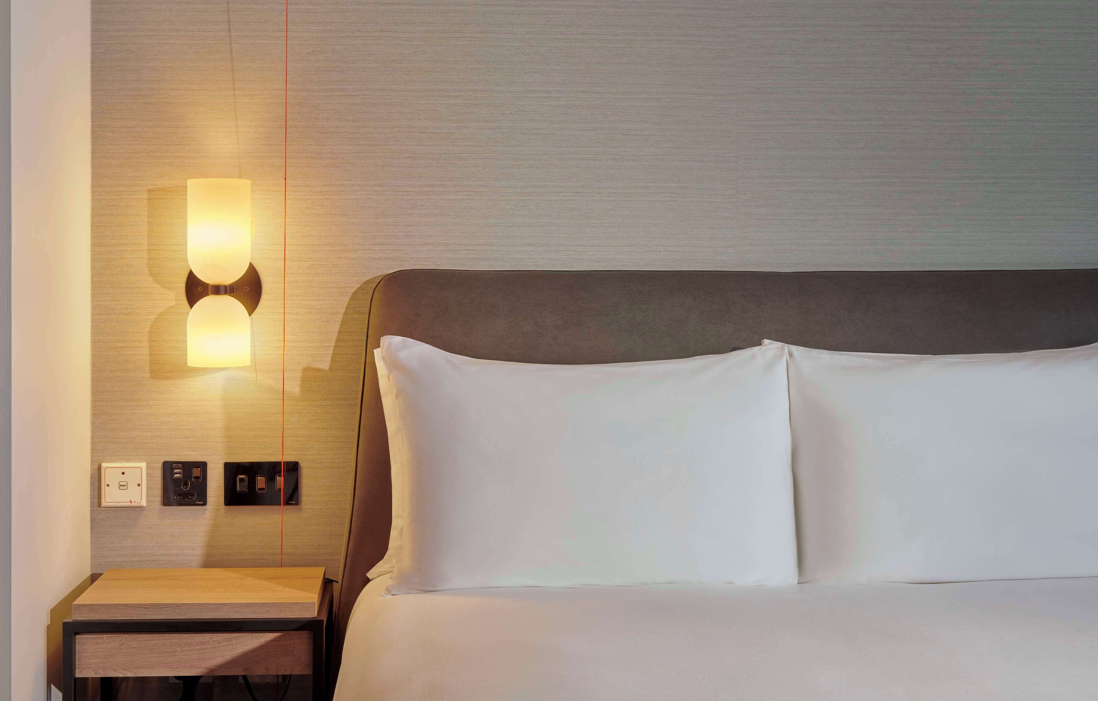 A hotel room with a bed, a bedside table, and a wall-mounted light fixture.