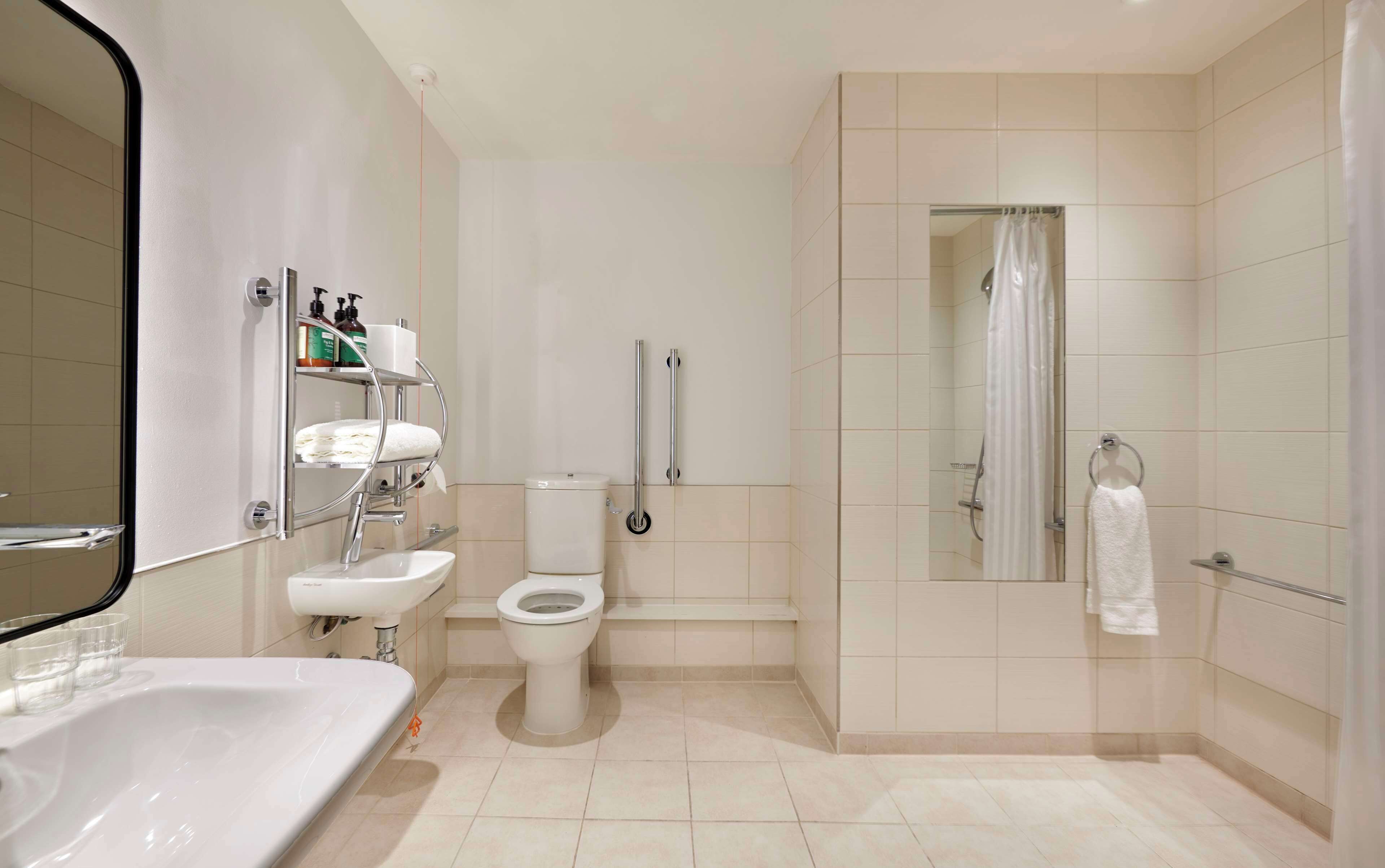 A modern bathroom with a white sink, toilet, and shower. There is a mirror, a shelf with toiletries, and a towel rack.