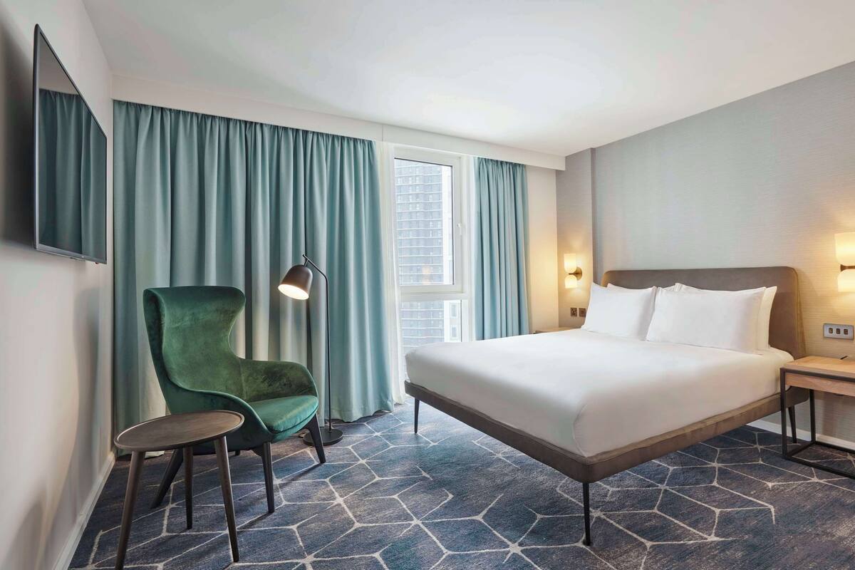 Ein Hotelzimmer mit einem Bett, einem grünen Sessel, einer Stehlampe und einem Nachttisch.