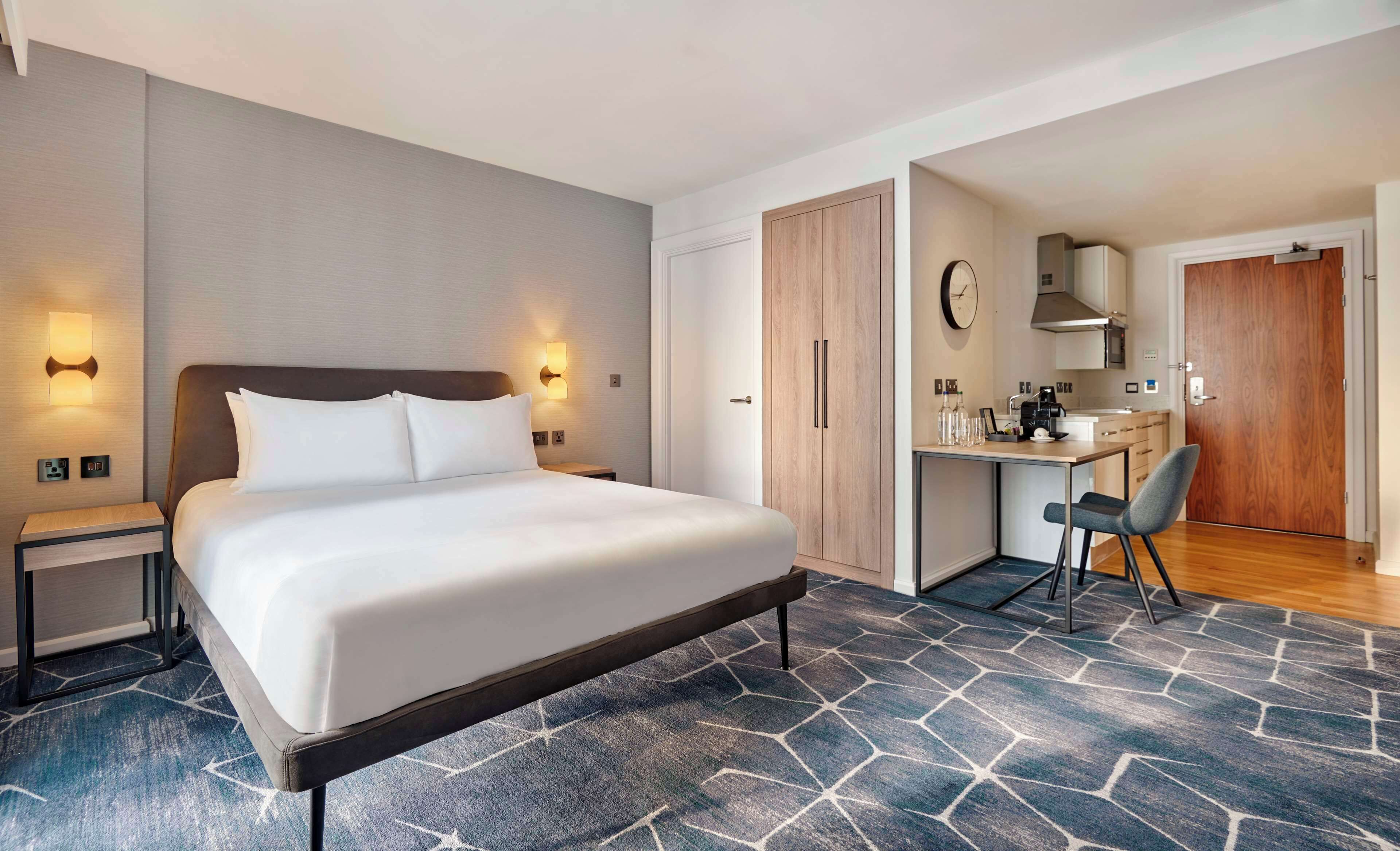 Une chambre d’hôtel moderne dotée d’un grand lit, de tables de chevet, d’un bureau avec une chaise, d’une armoire et d’une kitchenette.