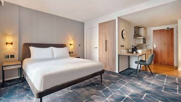 Une chambre d’hôtel moderne dotée d’un grand lit, de tables de chevet, d’un bureau avec une chaise, d’une armoire et d’une kitchenette.