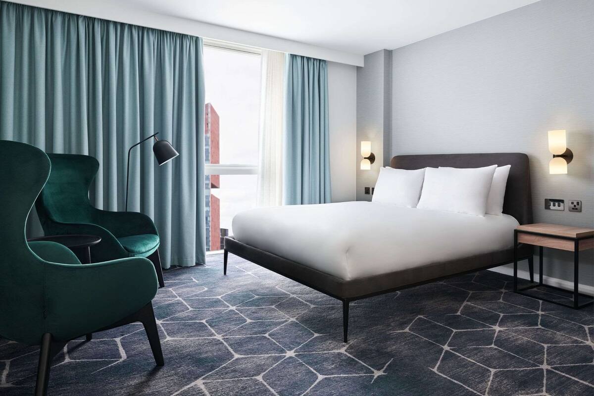 Ein Hotelzimmer mit einem großen Bett, einem grünen Sessel, einem Nachttisch mit Lampe und Blick auf Gebäude durch das Fenster.