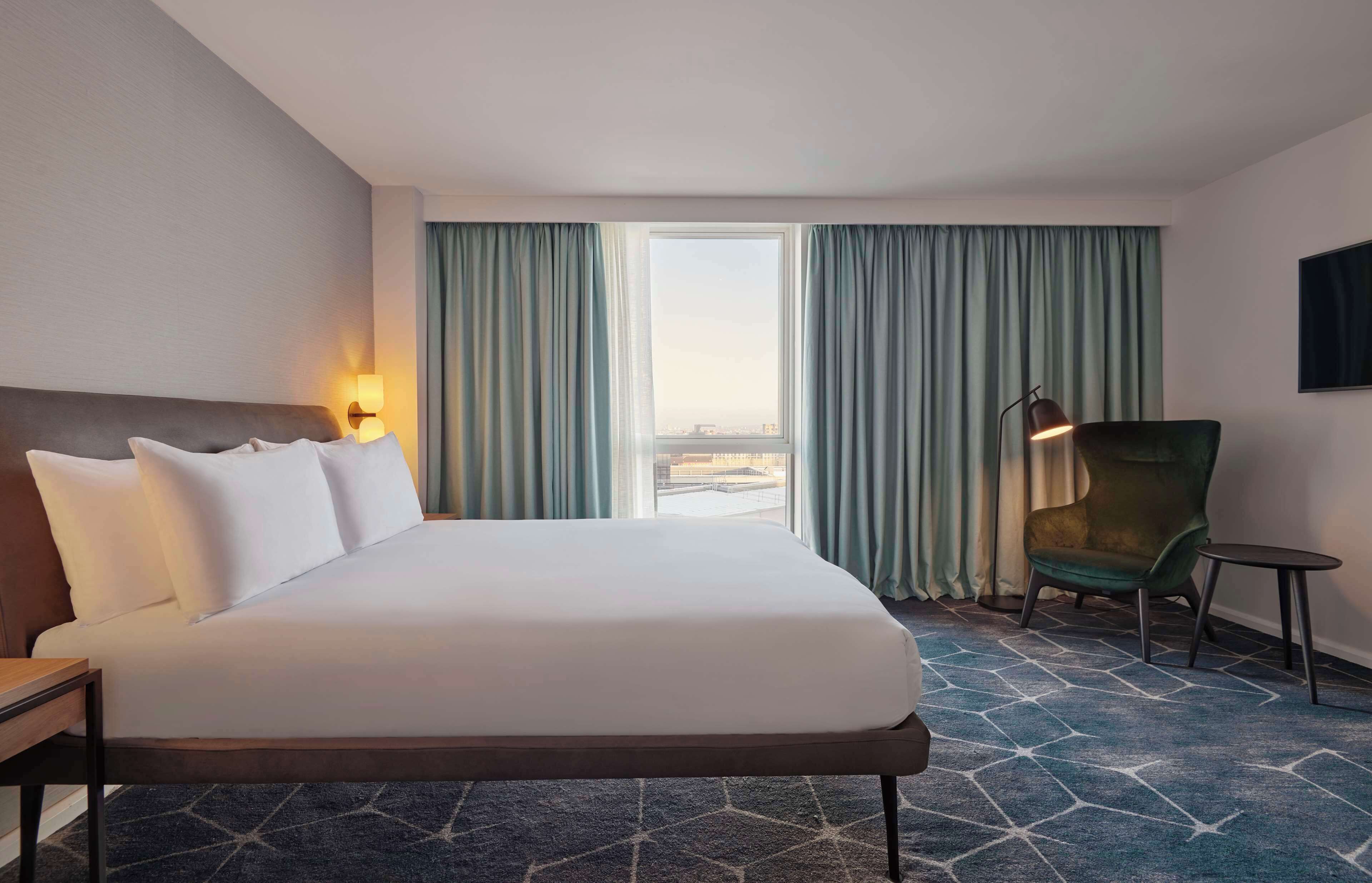 Ein Hotelzimmer mit einem großen Bett, einem Sessel, einem Schreibtisch und Blick auf die Stadt.