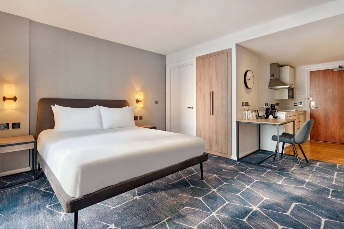 Ein Hotelzimmer mit Bett, Nachttischen, Schreibtisch, Stuhl und einer Küchenzeile.