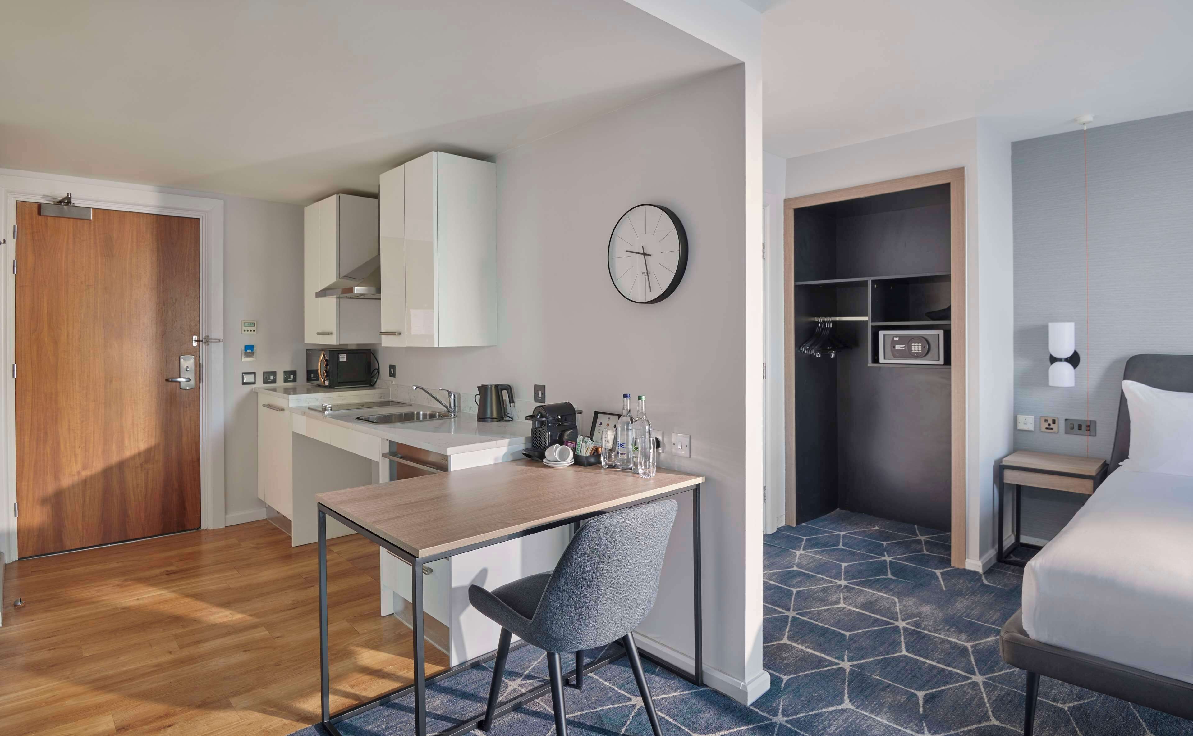 A compact hotel room with a kitchenette, a bed, a small table with a chair, and a clock on the wall.