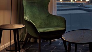 Una habitación moderna con un sillón verde, una lámpara de pie y una peque ña mesa redonda. Un ventanal grande ofrece vistas a un paisaje urbano nocturno.
