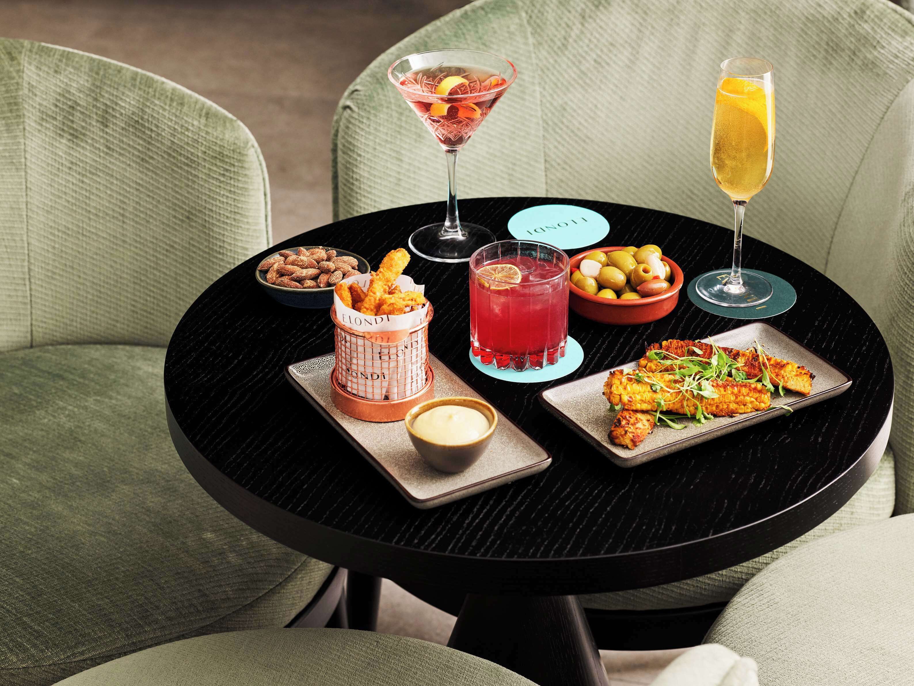 A table with a martini glass, a red cocktail, a glass of orange juice, a basket of fried chicken tenders, a bowl of olives, a plate of grilled chicken, and a small bowl of sauce.