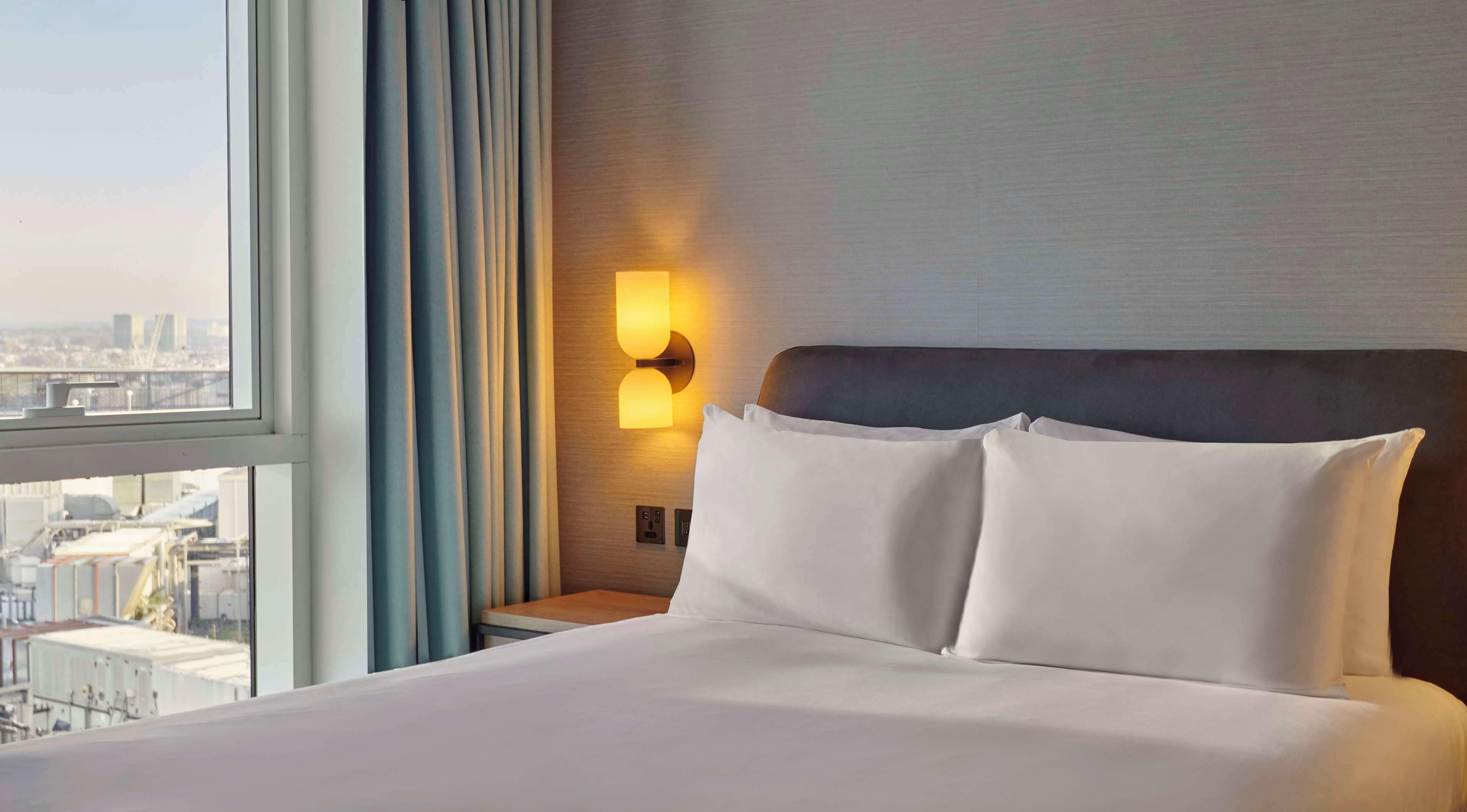 A hotel room with a large bed, a nightstand with a lamp, and a view of the city through the window.
