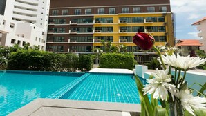 A swimming pool with a tiled deck, surrounded by greenery and a building with multiple balconies in the background. - Neo Hotel Pattaya (Pattaya)