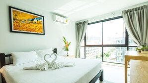 A hotel room with a large bed, a painting on the wall, a window with curtains, and a view of buildings outside. - Neo Hotel Pattaya (Pattaya)