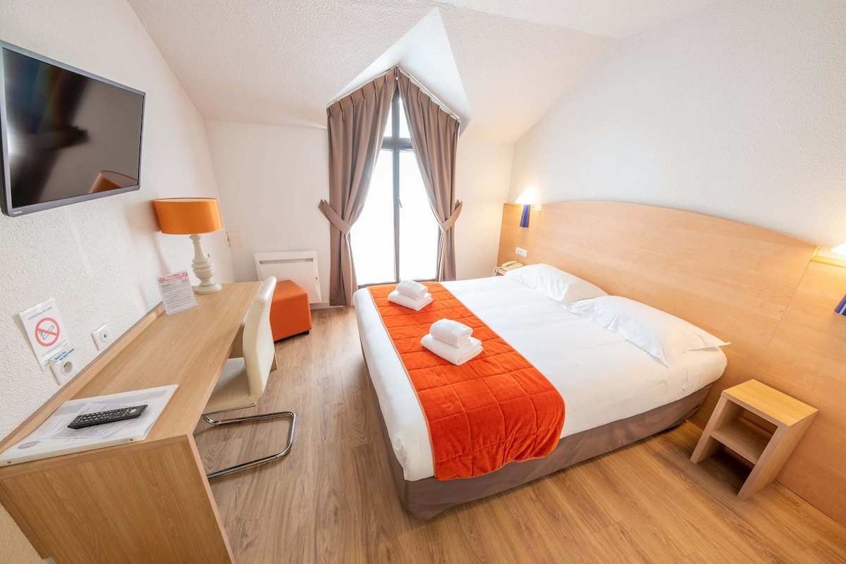A hotel room with a bed, a desk with a chair, a TV, and a window with curtains.