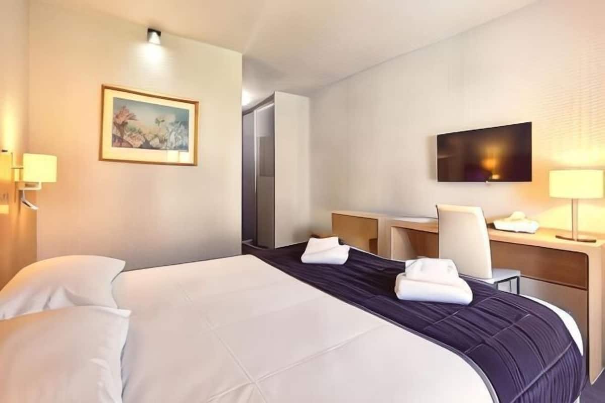 A hotel room with a bed, a desk, a chair, a television, and a painting on the wall.