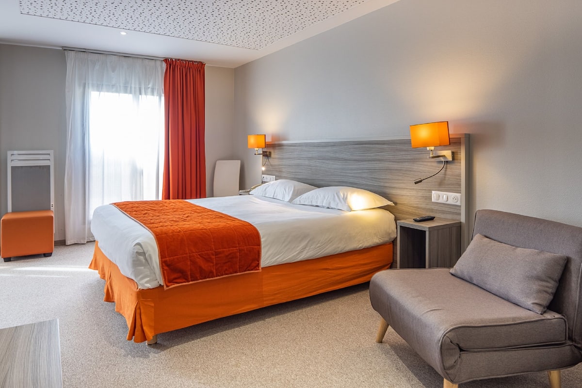 A hotel room with a bed, a chair, a nightstand, a lamp, and a window with curtains.