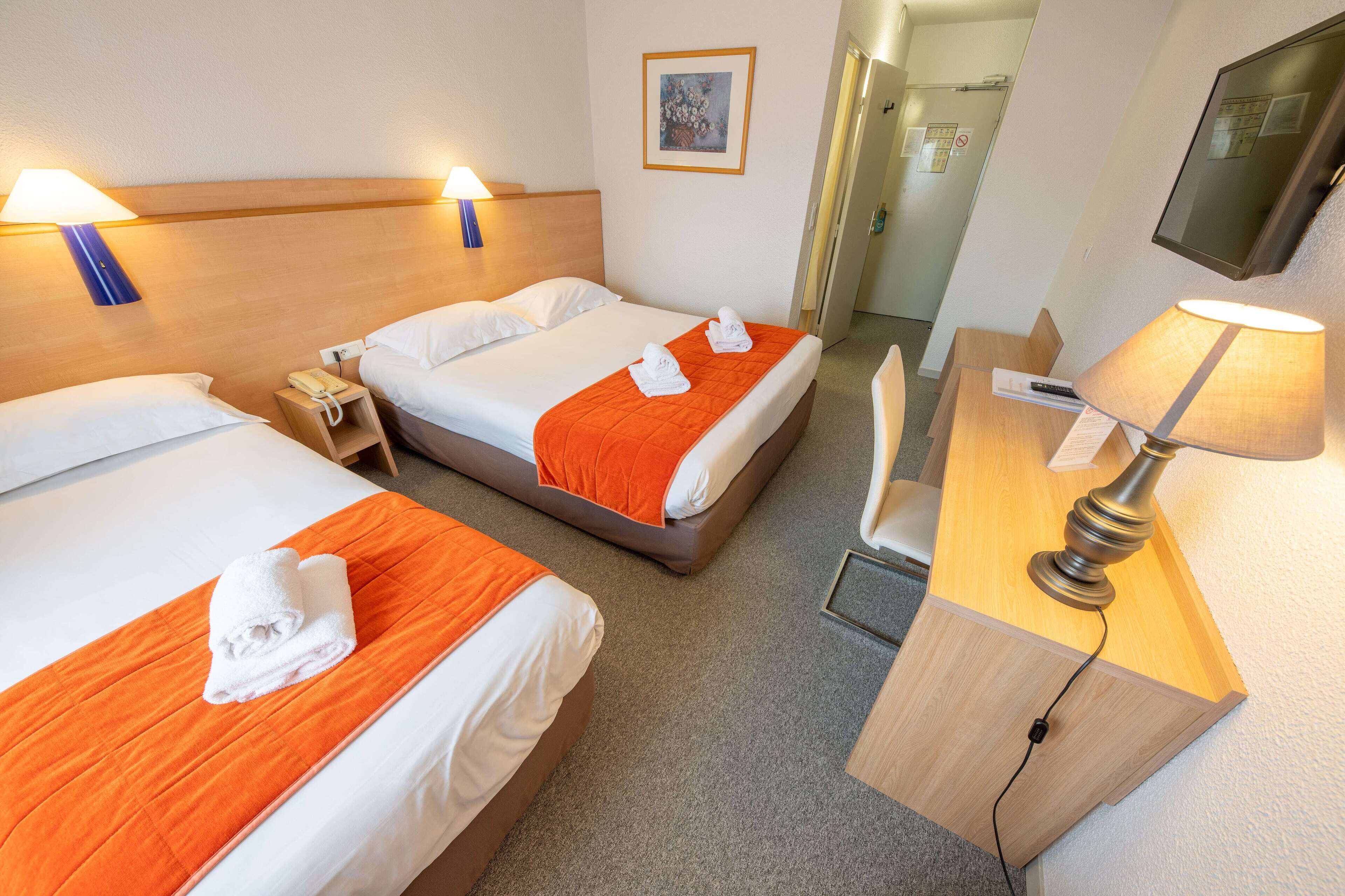 Une chambre d’hôtel avec deux lits, chacun recouvert d’une couette blanche et agrémenté de plaids orange. On y trouve des tables de chevet avec des lampes, ainsi qu’un petit bureau également doté d’une lampe.