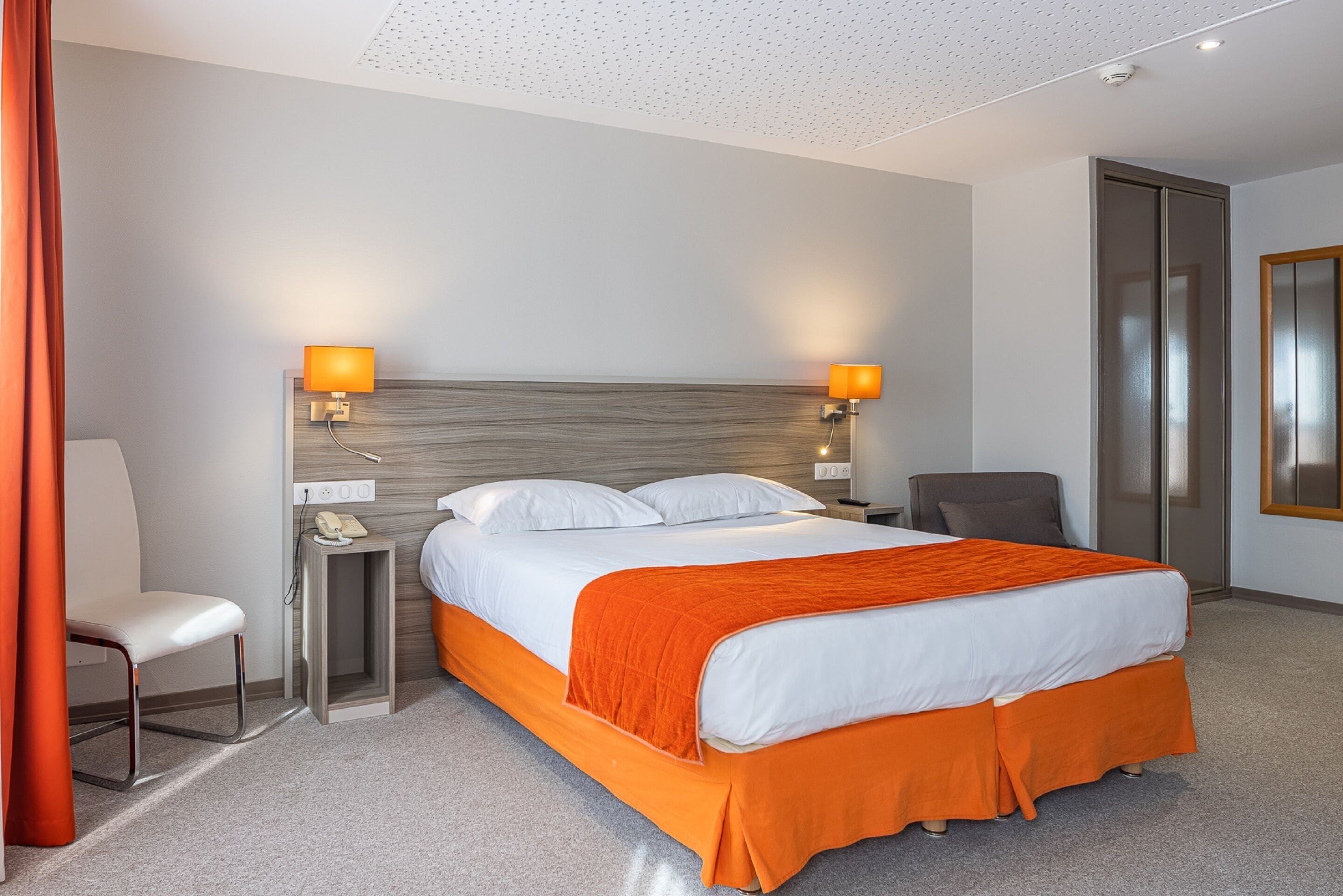 A hotel room with a bed, two bedside lamps, a chair, a desk with a phone, and a door.