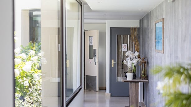 Un couloir moderne avec une grande fenêtre, une porte avec une pancarte et une petite table sur laquelle est posé un vase de fleurs.