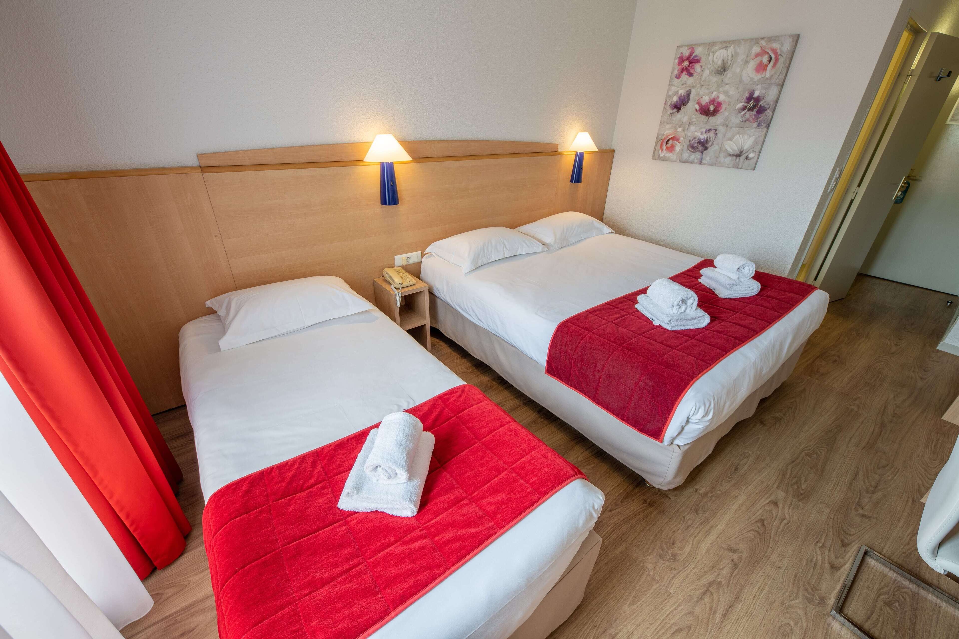 Hotellihuoneessa on kaksi sänkyä, joissa kummassakin on punainen peitto ja valkoiset pyyhkeet. Sängyn päädyn yläpuolella on seinään kiinnitetyt lamput, seinällä on taulu ja oikealla puolella ovi.