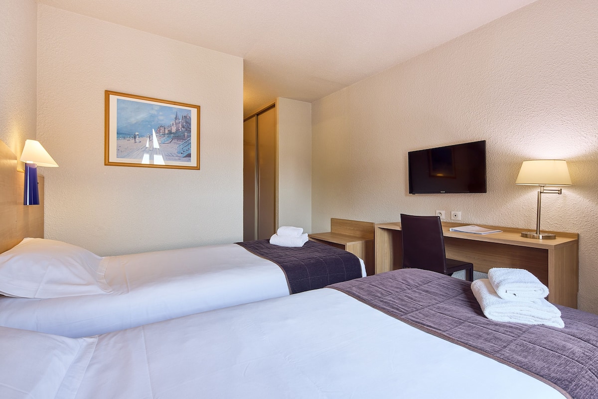 A hotel room with two beds, a desk, a chair, a TV, and a painting on the wall.