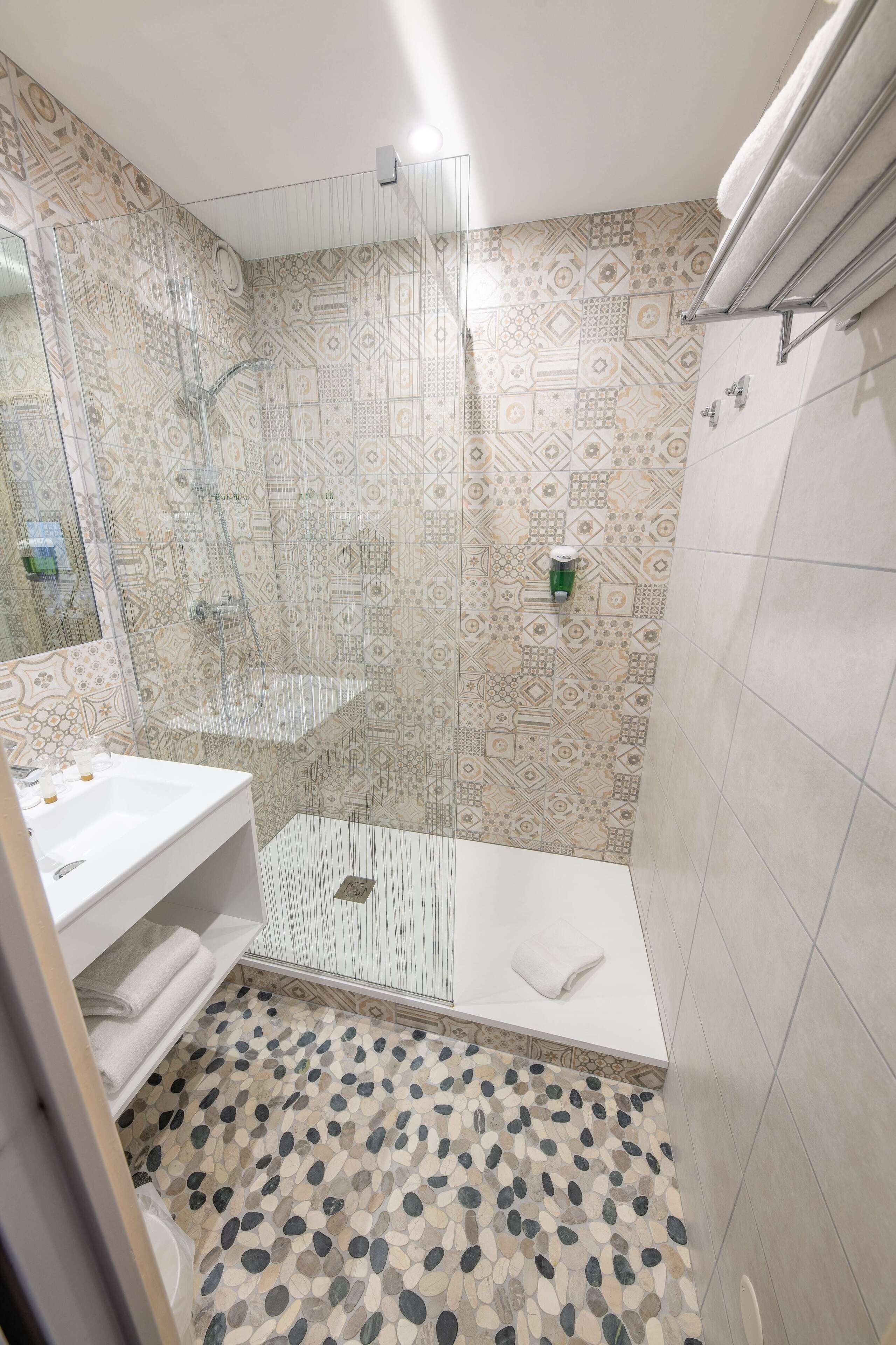 Een badkamer met een douchecabine van glas, betegelde wanden, een wit wastafelmeubel en een vloer van kleine kiezelstenen.