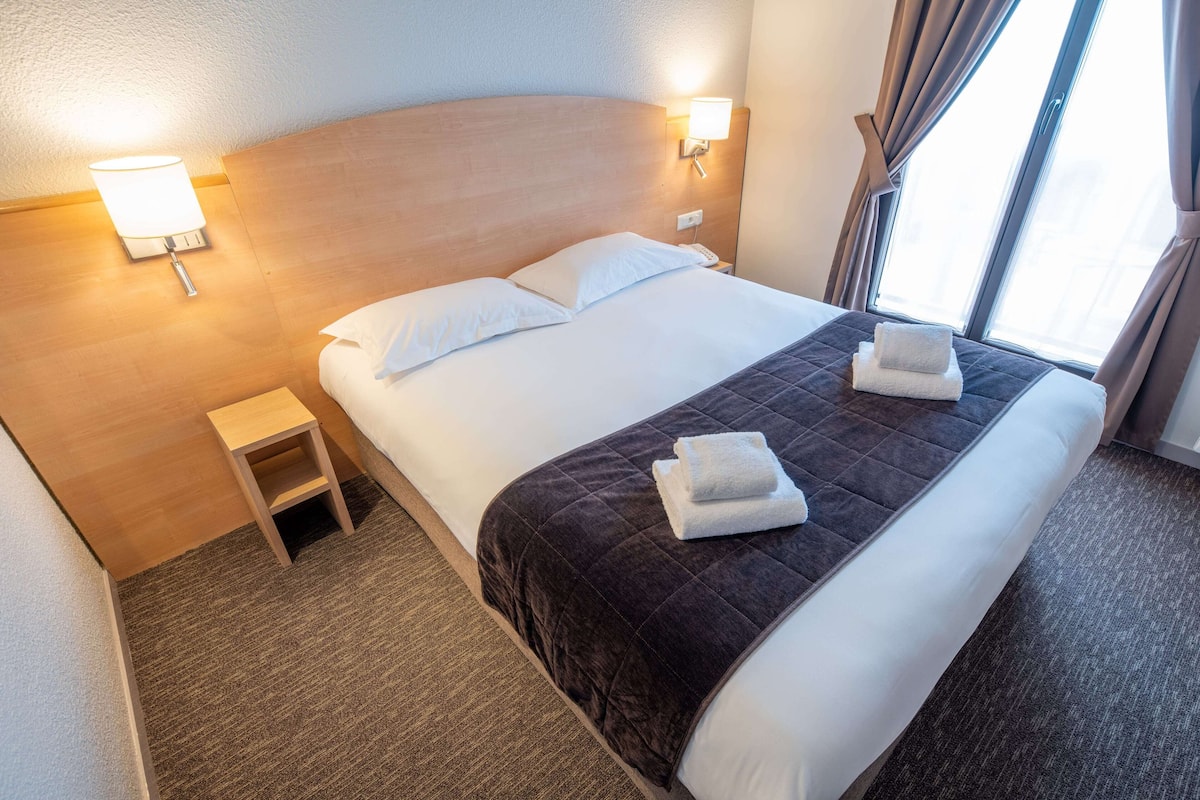 A neatly made bed with a dark quilt and two folded towels, a small bedside table, and two wall-mounted lamps.