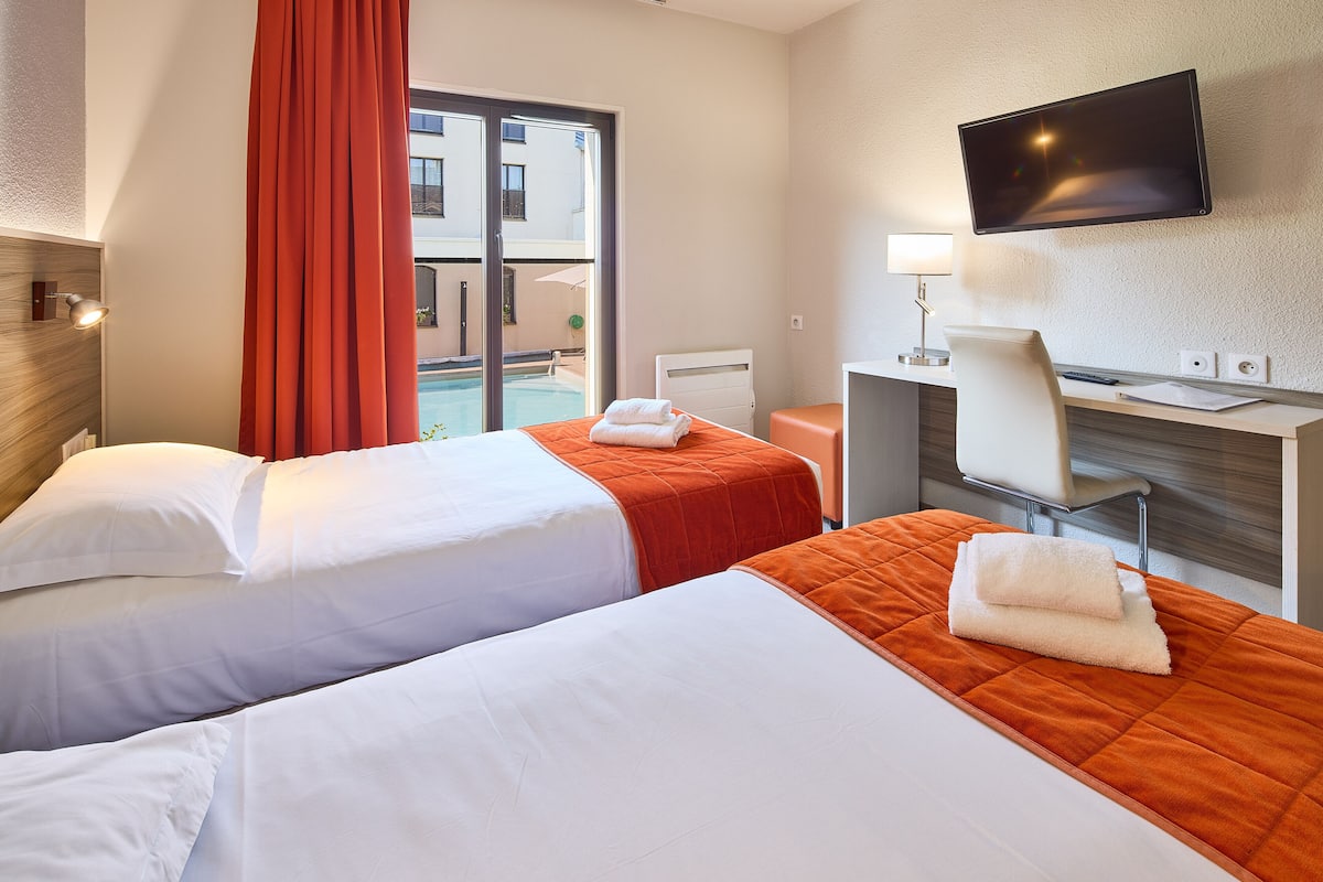 A hotel room with two beds, a desk, a chair, a TV, and a window with a view of buildings.