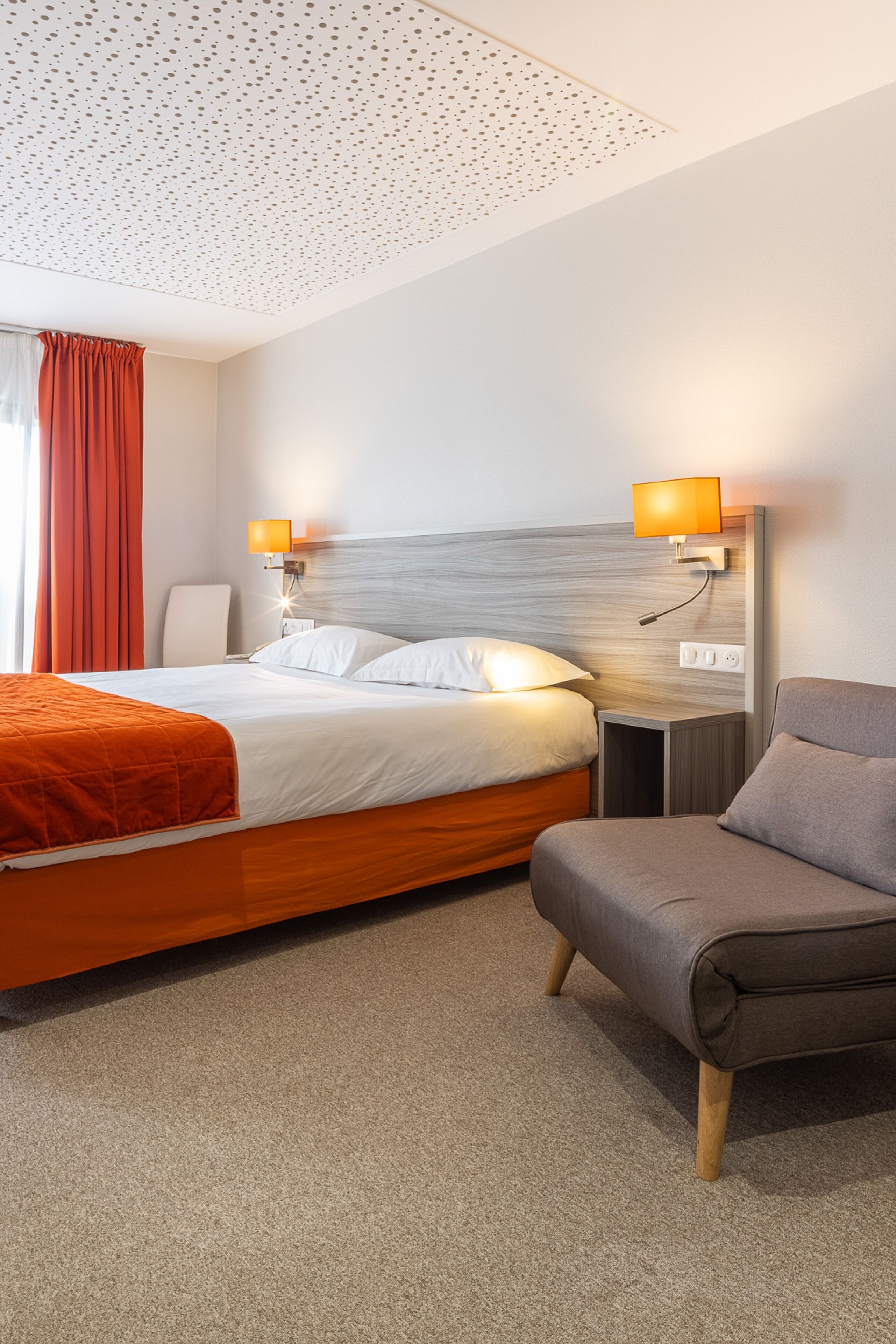 Une chambre d’hôtel avec un lit, deux lampes de chevet, une chaise et un sol recouvert de moquette.