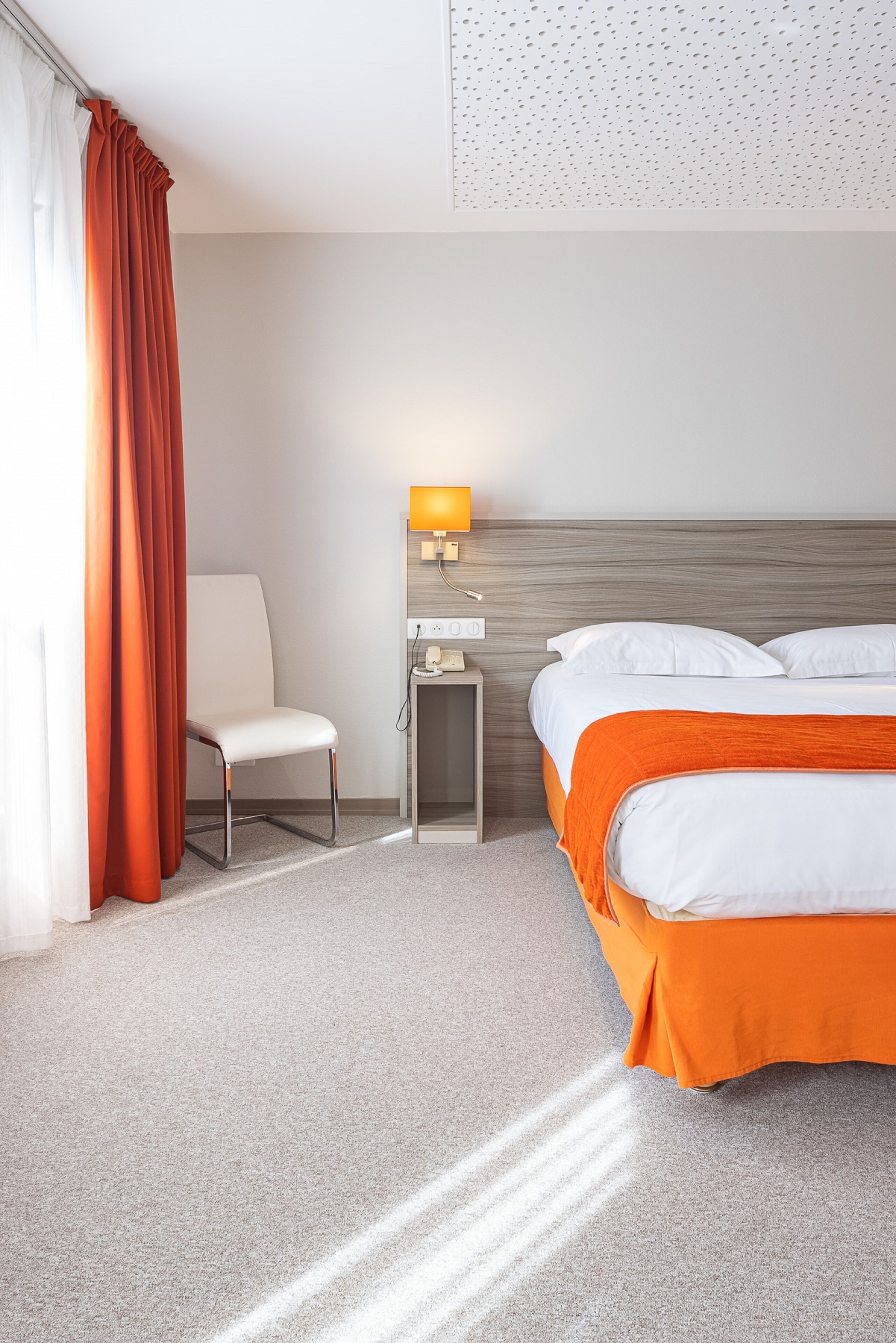 A hotel room with a bed, a chair, a nightstand with a lamp, and a wall with a perforated design.