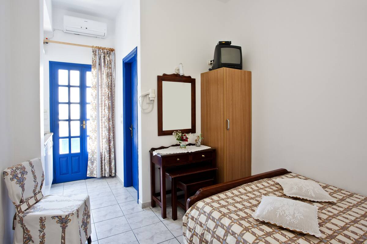 A bedroom with a blue door, a wooden wardrobe, a bed with a patterned bedspread, a wooden nightstand, and a chair with a floral cover.