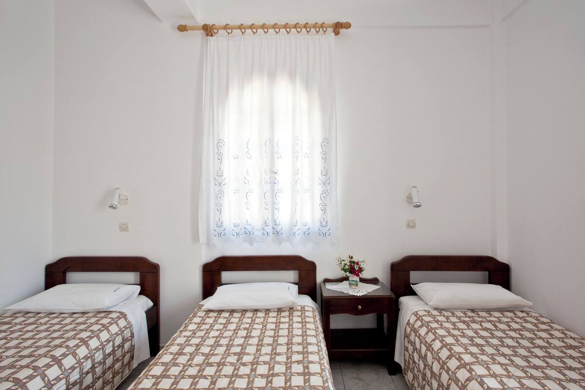 A room with three single beds, a small bedside table with a lamp, and a window with sheer curtains.