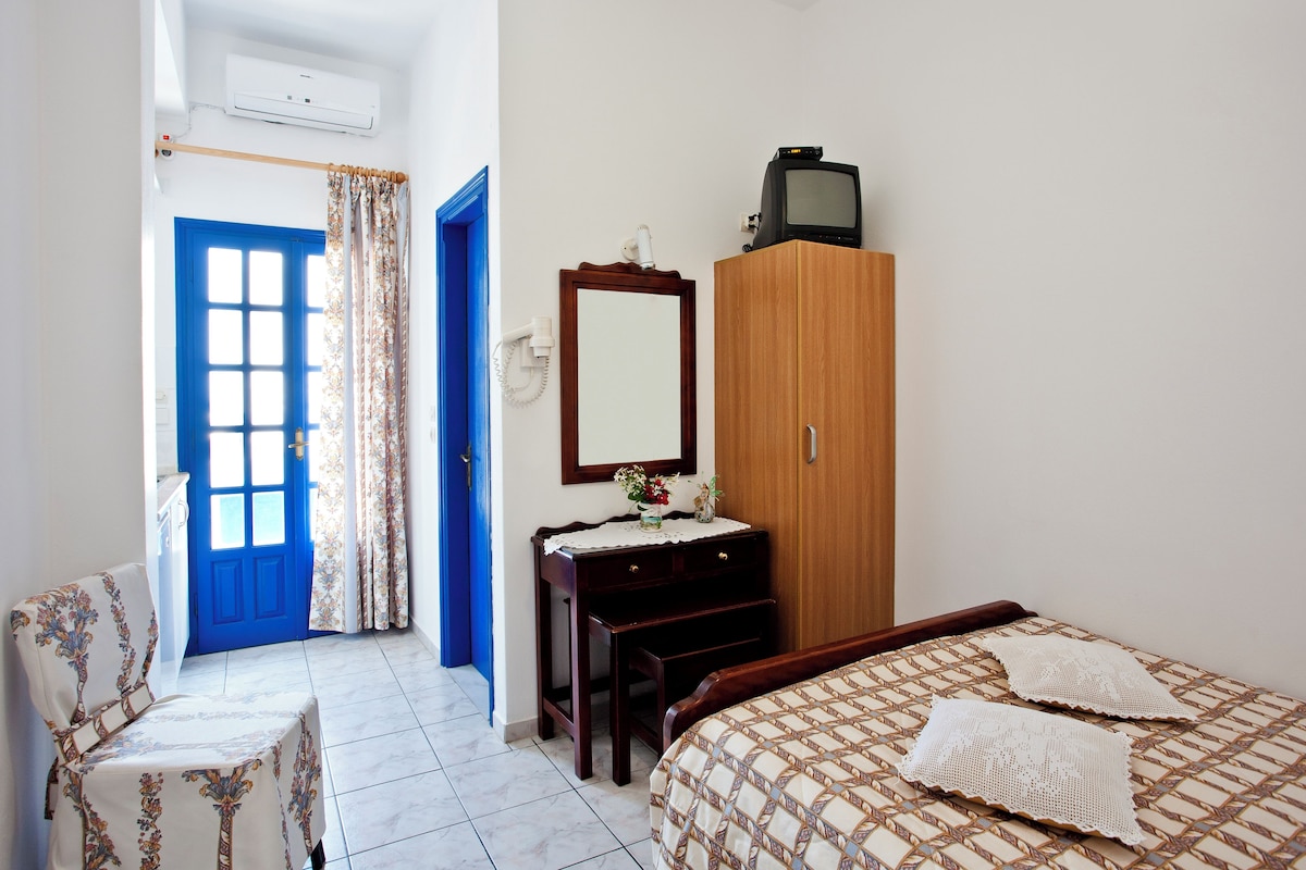 A bedroom with a blue door, a wooden wardrobe, a bed with a patterned bedspread, a wooden nightstand, and a chair with a floral cover.