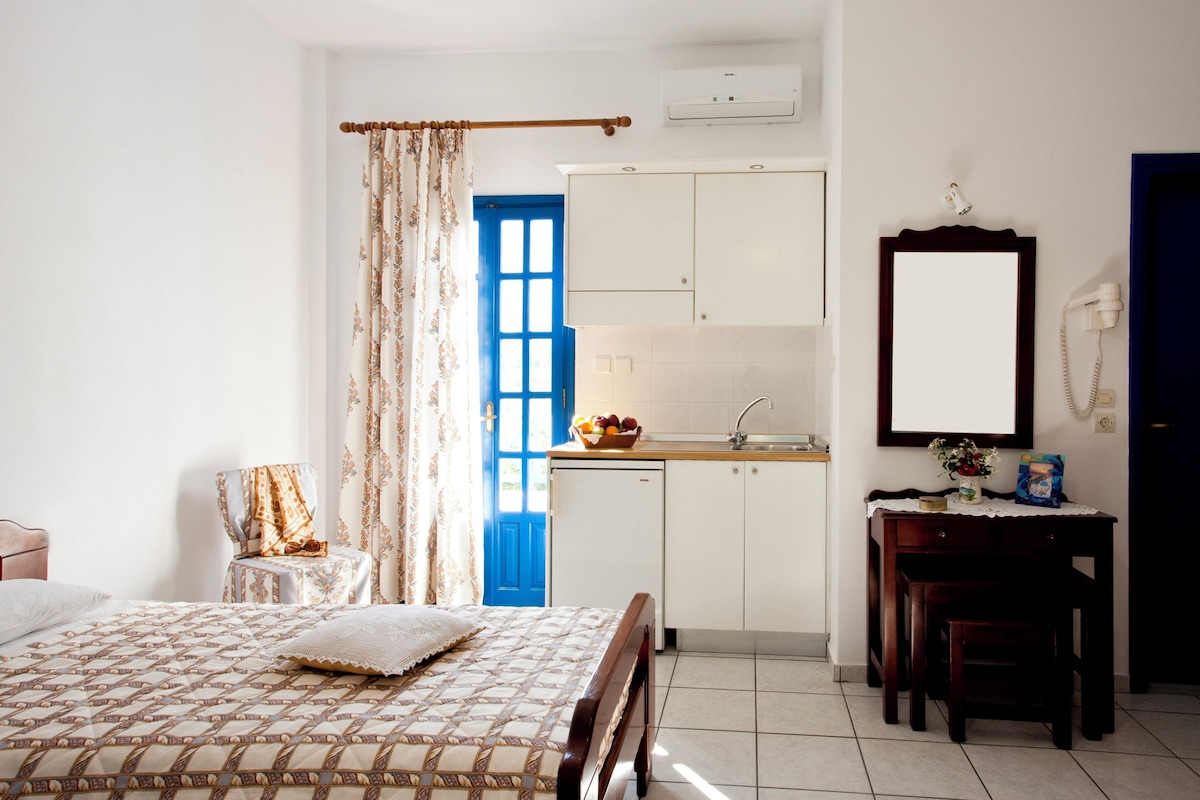 A bedroom with a bed, a chair, a small table, a desk, a mirror, a sink, and a blue door.