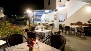 Un espace extérieur aménagé pour les repas, avec des tables et des chaises, une enseigne indiquant « EATOPIO ANCIENT THERA RESTAURANT », et un bâtiment à la façade blanche agrémentée de touches de bleu.