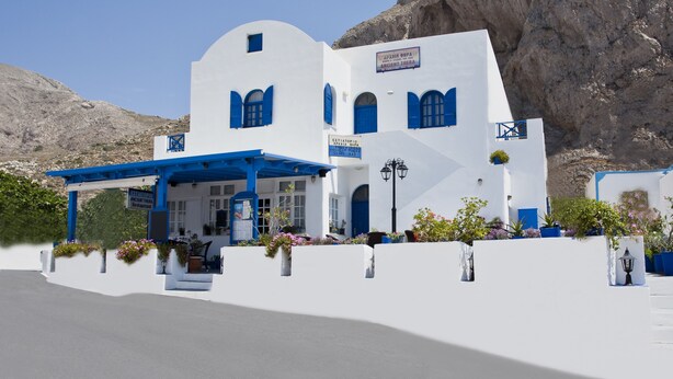 Un bâtiment blanc agrémenté de touches de bleu, une enseigne indiquant « Estia Topos, théâtre antique », et un paysage montagneux en arrière-plan.