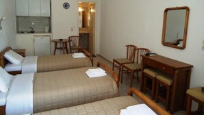 A hotel room with two beds, a desk, a chair, a small table, a mirror, and a kitchenette. - Pension Romantika (Mykonos)