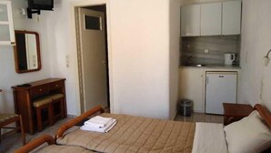 A compact room with a bed, a small desk, a mirror, a television, a kitchenette, and a door. - Pension Romantika (Mykonos)
