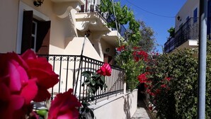 A narrow street with a white building, a black metal fence, and red flowers. - Mela Studios (Kalymnos)