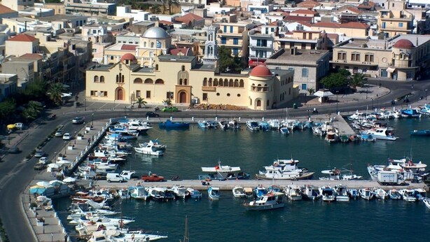 A coastal town with a marina filled with boats, a prominent yellow building with a dome, and densely packed residential buildings. - Mela Studios (Kalymnos)