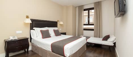Una habitación de hotel con una cama grande, una mesita de noche con lámpara, un televisor montado en la pared y una ventana con cortinas.
