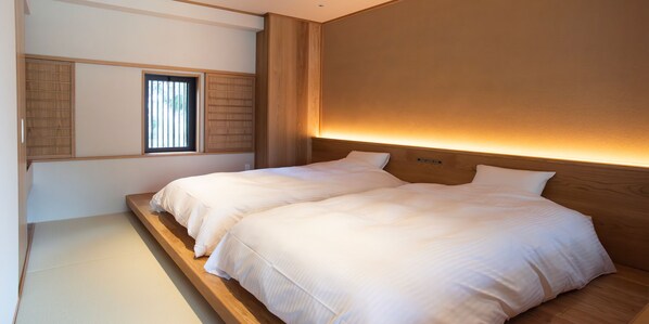 A bedroom with two beds, a wooden headboard, a window with blinds, and a wall-mounted light.