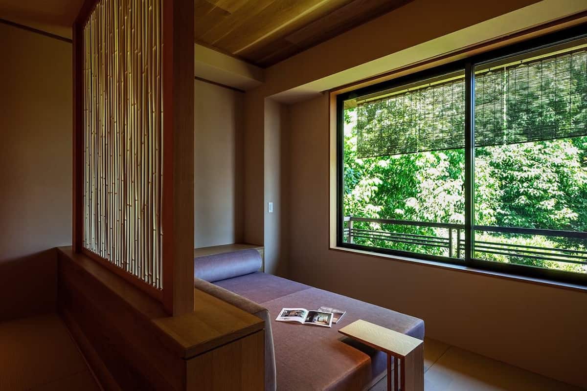 A room with tatami flooring, a low wooden bench, a cushioned seating area, and a large window offering a view of greenery outside.