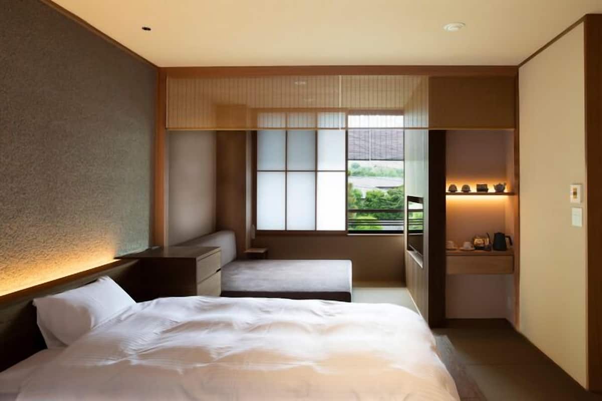 A hotel room with a bed, bedside tables, a small desk, a window with a view, and a shelf with items.