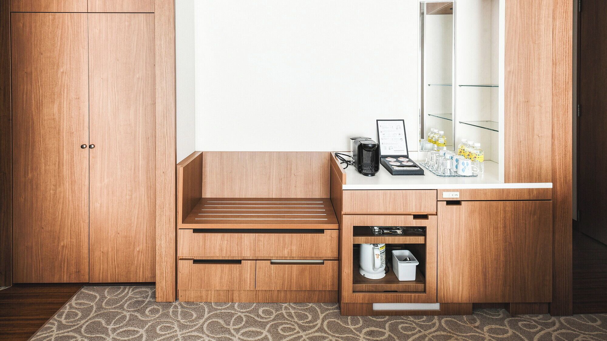 A compact bathroom with a wooden vanity, a sink, and a mirror. There is a small shelf with toiletries and a towel rack above the mirror.