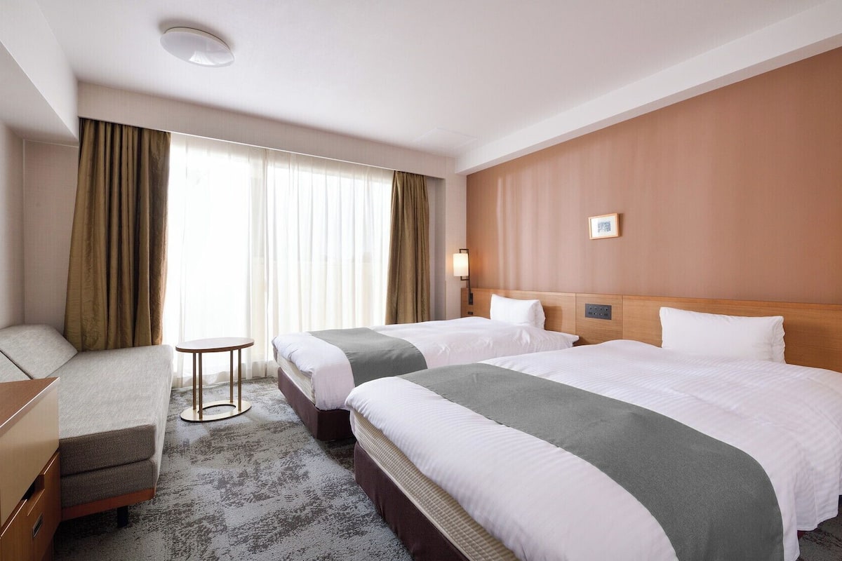 Une chambre d’hôtel avec deux lits, une petite table, un canapé et une fenêtre avec des rideaux.