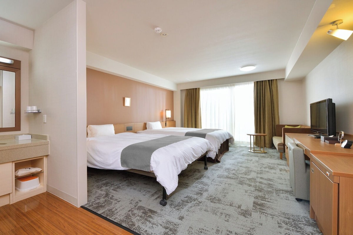 Une chambre d’hôtel avec deux lits, un bureau avec une télévision, une salle de bain avec un miroir et un lavabo, et une grande fenêtre avec des rideaux.