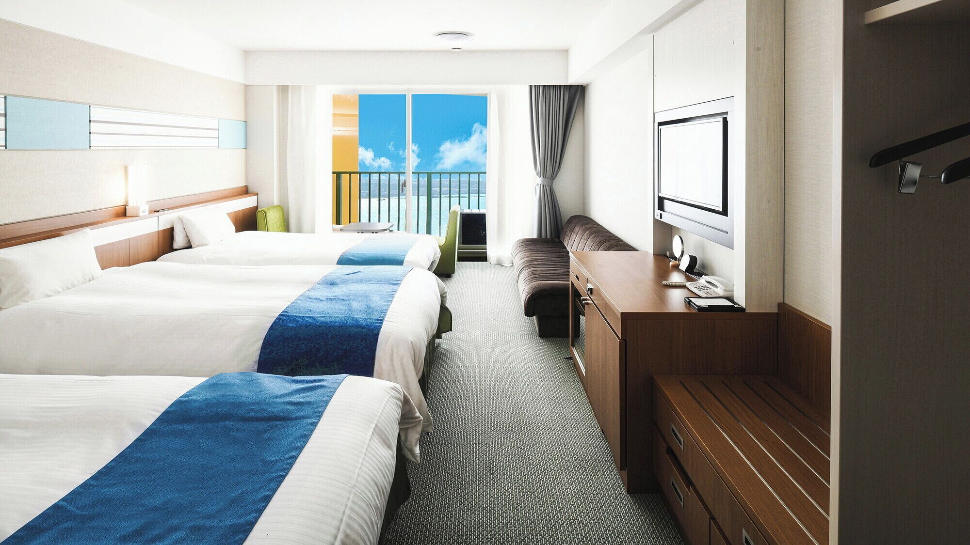 A hotel room with two beds, a desk, a chair, a TV, and a balcony with a view of the sea.