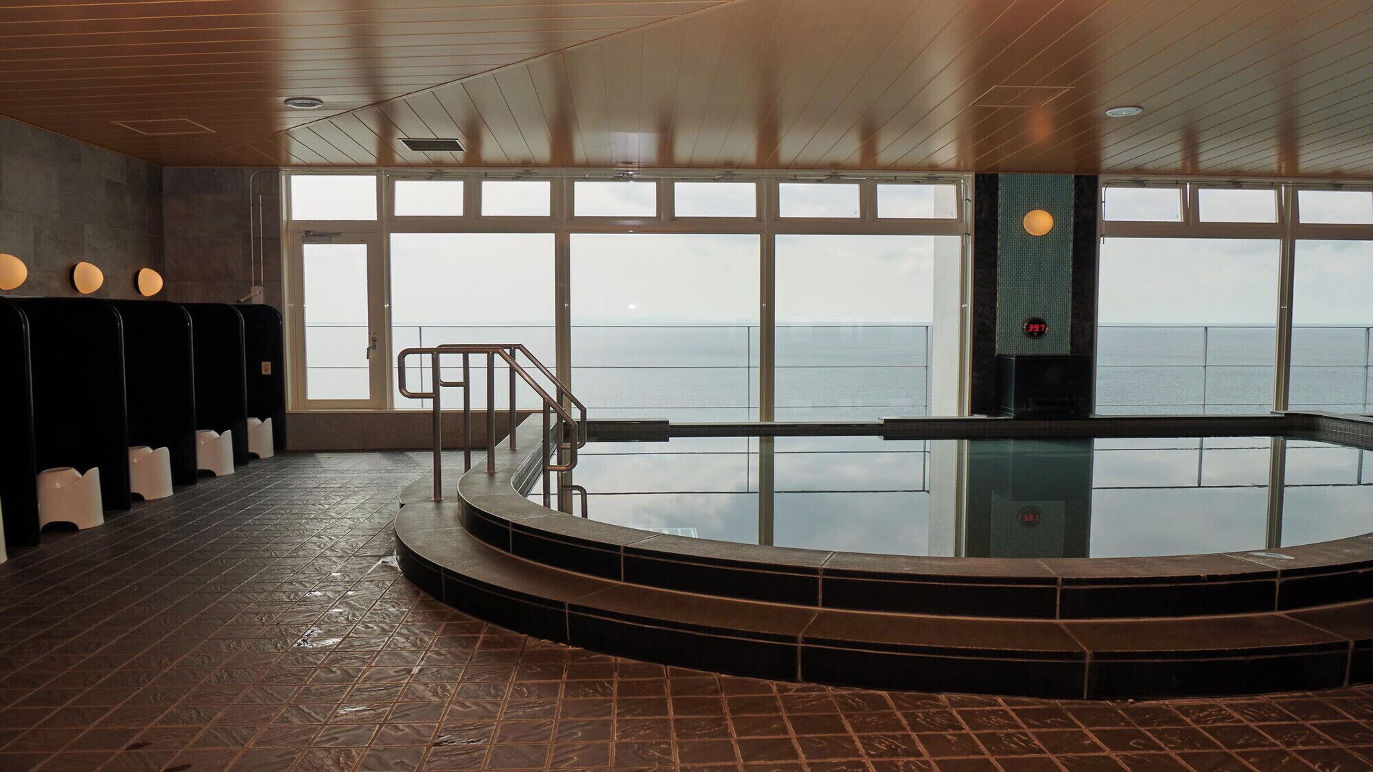 An indoor swimming pool with a view of the sea, a staircase leading to the pool, and a row of changing rooms with lights above them.