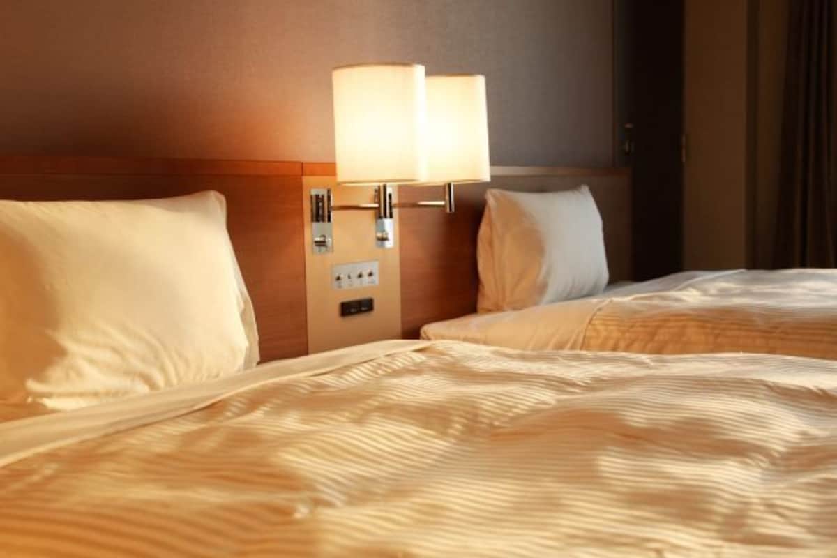 A hotel room with two beds, a nightstand with a lamp, and a wall-mounted control panel.