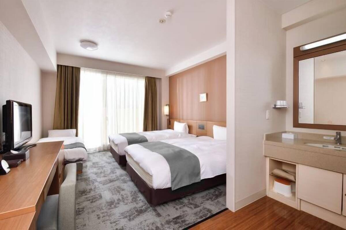 Une chambre d’hôtel avec deux lits, un bureau, une télévision, un grand miroir, une salle de bain avec un lavabo et un miroir, et une fenêtre avec des rideaux.